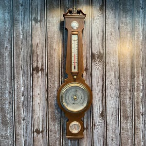 Peut inclure: Station météorologique en bois avec thermomètre, baromètre et hygromètre. Le thermomètre a un indicateur rouge. Le baromètre a un cadran circulaire avec des chiffres et une aiguille. L'hygromètre est un petit cadran circulaire en bas. Le bois est de couleur marron chaud.