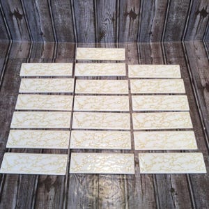 May include: A collection of rectangular white tiles with a gold, organic pattern. The tiles are arranged on a wooden surface, showcasing their decorative design. The tiles are likely for home decor or renovation projects.
