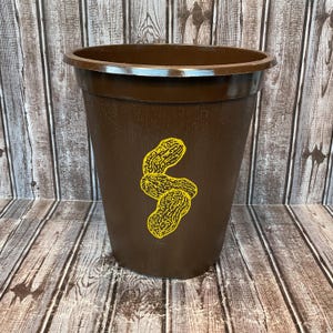 May include: A brown plastic planter with a wide rim and a tapered body. The planter features a yellow graphic of three peanuts. The background is a wooden surface.