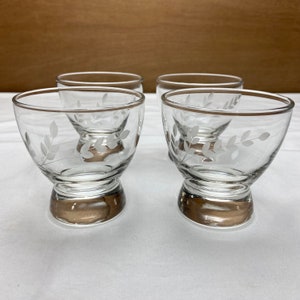 May include: Set of four clear glass drinking glasses with a white etched leaf design and a gold rim. The glasses have a short stem and a rounded base.