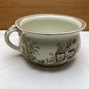 May include: Cream-colored antique chamber pot with a brown floral and landscape design. The pot has a handle and a decorative rim. The design includes flowers, foliage, and architectural elements.