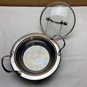 May include: A stainless steel cooking pot with two handles and a clear glass lid. The lid is off to the side, revealing the interior of the pot. The pot has a shiny, reflective surface, and the lid has a stainless steel handle.