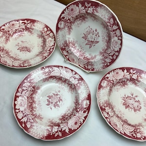 May include: Four antique ceramic plates with a floral design in burgundy and white. The plates feature a central floral bouquet and a border of flowers and foliage. The plates have a scalloped edge.