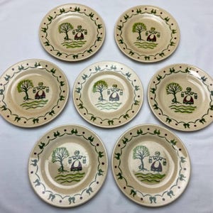 May include: Set of seven ceramic plates with a cream-colored base. Each plate features a hand-painted scene of a woman carrying buckets near a tree, with a green and red border. The plates are likely vintage or antique.