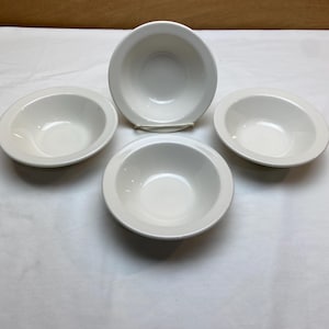 May include: Four white ceramic bowls with a rounded shape and a wide rim. The bowls are arranged on a white surface, with one bowl slightly elevated. The bowls are empty and appear clean.