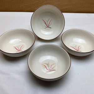 May include: A set of four off-white ceramic bowls, each featuring a simple, stylized pink and brown leaf-like graphic at the center. The bowls have a smooth, rounded shape and a thin, dark brown rim around the top edge of each piece.