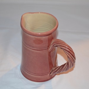 May include: A pink ceramic pitcher with a twisted handle. The pitcher has a cream-colored interior.