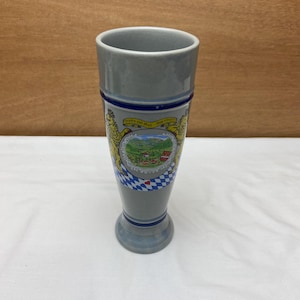 May include: A tall, gray ceramic beer stein with a blue and white checkered pattern and a crest featuring two lions and the text "Gruber und Mail, vom Jahre 1870".
