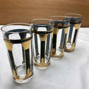 May include: Set of four clear drinking glasses with a decorative design. Each glass features a dark teal band at the top and bottom, accented with gold detailing. Vertical black and gold columns are painted on the glass, creating an elegant aesthetic.
