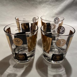 May include: Set of four clear glass tumblers with gold and black geometric designs. Each glass features a silver coin design with the text "United States of America" and "2 1/2 Dollars".