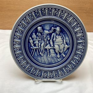 May include: A blue and white ceramic plate with a raised design of two men in a scene from a tavern. The plate has a decorative border.