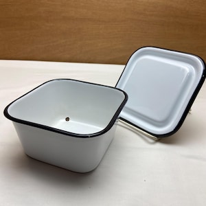 May include: A square white enamel storage container with a matching lid. The container and lid both feature a smooth white finish with a thin black rim along the edges. A small dark spot is visible on the interior base of the open container.