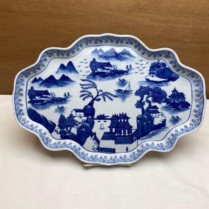 May include: A white porcelain serving platter with a blue landscape design. The platter has a scalloped edge with a detailed border. The blue design features mountains, buildings, trees, and water elements.