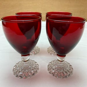 May include: Set of four ruby red wine glasses with clear, textured bases. The glasses have a classic shape and are perfect for serving wine or other beverages.