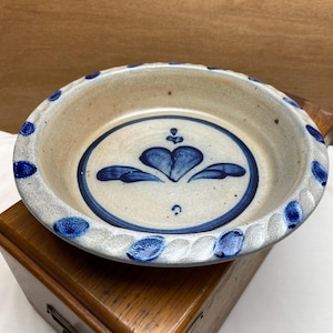 May include: A round, stoneware pie dish with a cream-colored interior and a blue heart design in the center. The rim features a series of blue dots and a textured edge. The dish is approximately 9 inches in diameter.