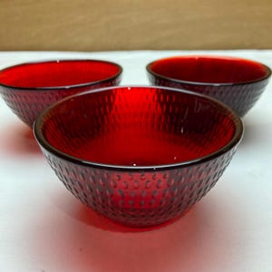 May include: Three ruby red glass bowls with a textured, dimpled surface. The bowls are arranged in a group, with the closest bowl in sharp focus. The bowls have a classic, rounded shape and are suitable for serving.