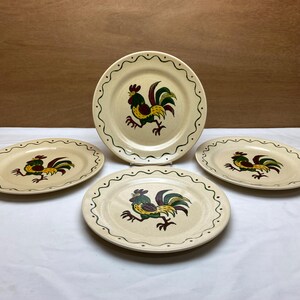 May include: Four ceramic plates with a rooster design. The plates have a cream-colored background with a scalloped edge. The rooster is depicted in green, yellow, and burgundy. The plates are arranged on a white surface.