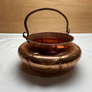 May include: A hammered copper pot with a curved handle. The pot has a rounded bottom and a wide, flared rim. The pot is made of copper and has a rustic, antique look.