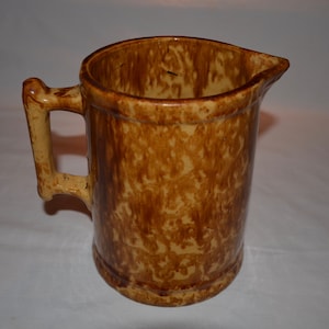 May include: A brown and yellow glazed ceramic pitcher with a handle. The pitcher has a mottled brown and yellow glaze.