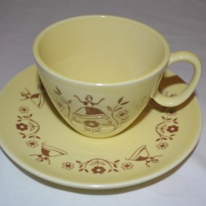 May include: A yellow teacup and saucer set with a brown floral pattern. The pattern features stylized figures and flowers.