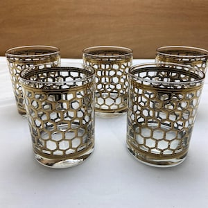 May include: Five clear drinking glasses with a gold honeycomb pattern. The glasses have a gold rim and base, and the honeycomb design covers the body of the glass. The glasses are arranged in a group, showcasing their design.