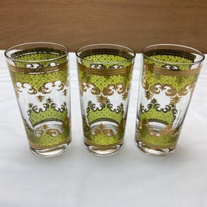 May include: Three clear drinking glasses with a green and gold decorative pattern. The glasses have a tall, slender shape and a gold rim. The green pattern includes a diamond-shaped design and ornate gold scrollwork.
