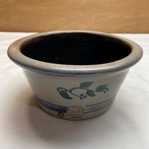 May include: A round stoneware bowl with a beige exterior and a dark brown interior. The exterior features a blue horizontal band, a green leaf and berry vine motif, and a small embossed rectangular maker's mark near the base rim.