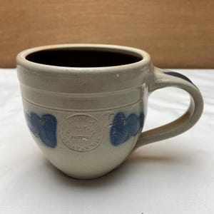 May include: A stoneware mug with a cream-colored base and a dark brown interior. The mug features blue heart-shaped designs and the embossed text "SHADOWLAWN STONEWARE POTTERY DELAVAN WIS". The handle is a matching cream color.