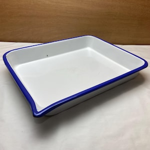 May include: A rectangular white enamel baking pan with a bright blue rim. The pan has a slightly curved edge and is empty. The background is a light brown wooden surface.