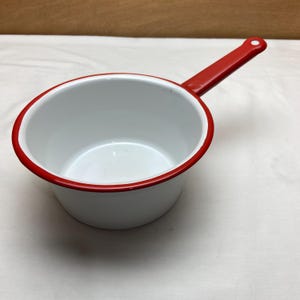 May include: A white enamel saucepan with a bright red rim and a matching red handle. The interior of the pan is smooth and white, while the exterior features a clean white finish. The handle includes a small hole at the end for hanging storage.