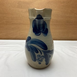 May include: A blue and white stoneware pitcher with a floral design. The pitcher has a wide mouth and a narrow neck. It is decorated with blue flowers and leaves on a white background. The pitcher is signed "Handmade in USA".