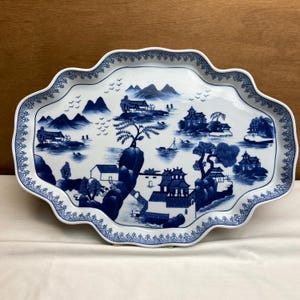 May include: An oval-shaped ceramic serving platter with a scalloped edge, featuring a blue and white transferware pattern of a landscape with mountains, pagodas, trees, and small boats on water, finished with a geometric border pattern.