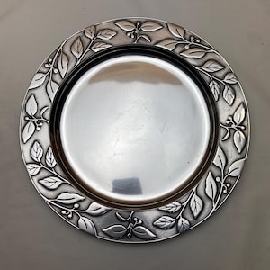 May include: A silver-colored plate with a detailed leaf and berry design around the rim. The plate has a smooth, reflective surface and a slightly raised edge. The leaf pattern adds a decorative touch to this serving dish.
