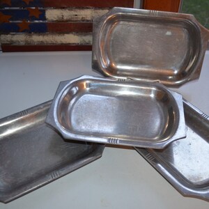 May include: Four vintage, rectangular metal trays with a silver finish. Each tray has a slightly raised, oval interior and decorative corner accents. The trays are arranged on a white surface, with a glimpse of a rustic American flag in the background.