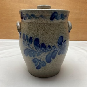 May include: A stoneware crock with a lid, featuring blue floral designs. The gray-beige pot has two handles and a matching lid with a small knob. The blue floral pattern decorates the body of the crock.