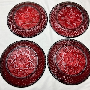 May include: Four ruby red glass plates with intricate starburst and geometric designs. The plates have a textured, ridged border and a central floral pattern. These decorative plates are ideal for serving or display.