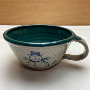May include: A ceramic soup mug with a speckled off-white exterior and a deep teal interior. The outer surface features a hand-painted blue line drawing of a snowman wearing a hat, surrounded by small blue dots and simple leaf-like accents.