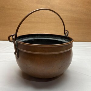May include: A copper cauldron with a curved handle. The pot has a rounded body and a dark interior. The handle is attached to the sides of the pot with rivets. The cauldron is likely used for cooking or decorative purposes.