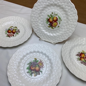 May include: Set of four white ceramic plates with a raised embossed design. Each plate features a central floral motif with fruit, including pears, grapes, and plums.