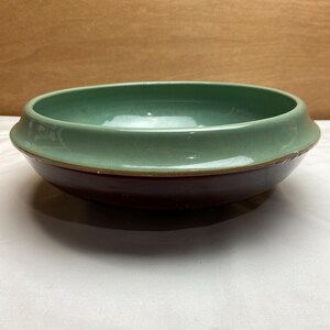 May include: A ceramic bowl with a wide, shallow design. The upper portion is a muted green, while the lower section is a deep burgundy. A thin, gold-colored band separates the two colors. The bowl sits on a white surface.