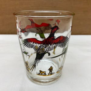May include: Clear glass tumbler featuring a vibrant illustration of a pheasant in flight, with red, black, and white plumage. The design includes additional birds and a hunting scene at the base. A vintage-style drinking glass.