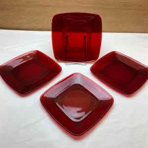 May include: A set of four ruby red glass plates. Three are diamond-shaped, and one is square. Each plate features a central circular design with radiating lines. The plates are displayed on a white surface.