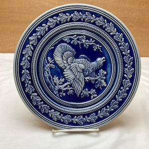 May include: A blue and white ceramic plate with a raised design of a bird on a branch. The plate is decorated with a border of oak leaves and acorns.