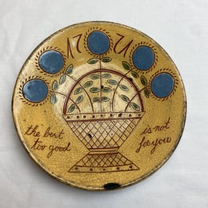 May include: A round yellow ceramic plate with a crackled glaze finish. It features a central illustration of a basket containing green leaves and four blue circular flowers. The text 1724 is at the top, and the text the best too good is not for you is written in brown script on the sides.