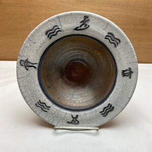 May include: A white ceramic bowl with a brown glaze inside. The bowl has a black rim and a design of waves, turtles, and people surfing around the rim.