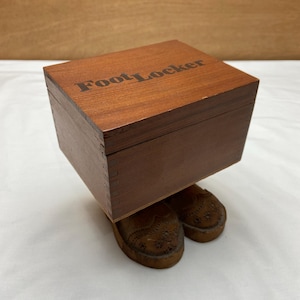 May include: A wooden box with the text "Foot Locker" on top, resting on a pair of brown, carved shoe forms. The box and shoes are a warm brown color, suggesting a vintage or handcrafted item.