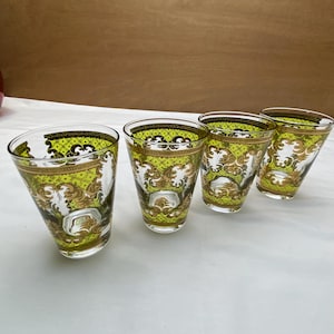May include: Four clear glass tumblers with a decorative design. The glasses feature a gold and green pattern with ornate detailing. The tumblers are arranged in a row, showcasing their elegant design.