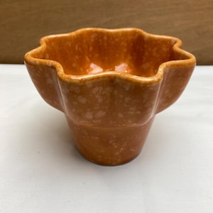 May include: An orange ceramic bowl with a unique, scalloped rim. The bowl has a speckled texture and a flower-like shape. The exterior is a solid orange color, while the interior is a lighter shade. This decorative bowl is suitable for serving or display.