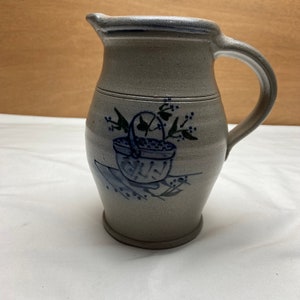 May include: A gray stoneware pitcher with a blue glaze and a hand-painted design of a basket of flowers. The pitcher has a wide mouth and a handle.
