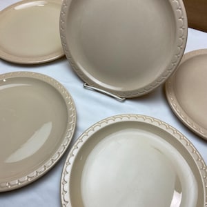 May include: A collection of beige ceramic plates. The plates have a smooth, slightly glossy surface and a decorative scalloped edge. The plates are arranged on a white surface, with some stacked and others lying flat.